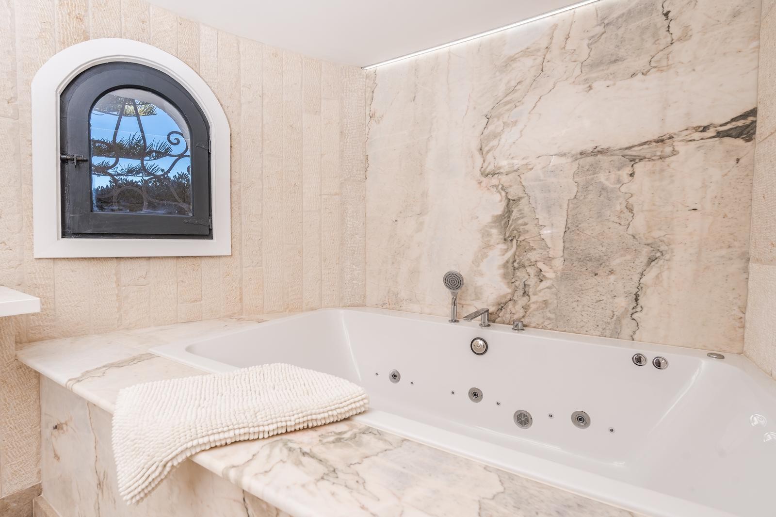 Eingebaute Whirlpool-Badewanne mit edler Marmorverkleidung und Natursteinwänden. Ein mediterranes Rundbogenfenster und LED-Lichtleisten setzen Akzente.