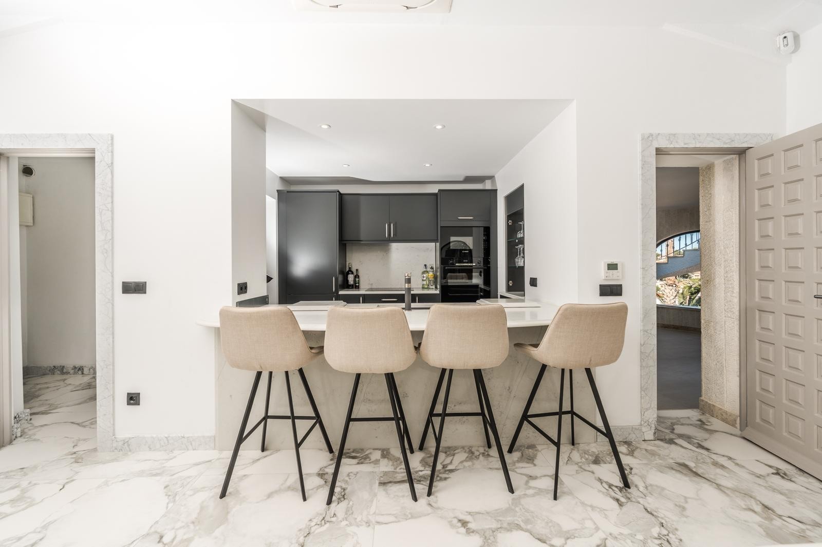Halboffene Küche mit Frühstücksbar, vier Polsterhockern und dunklen Schränken. Hochwertiger Marmorboden und Türrahmen aus Naturstein prägen das moderne Design.