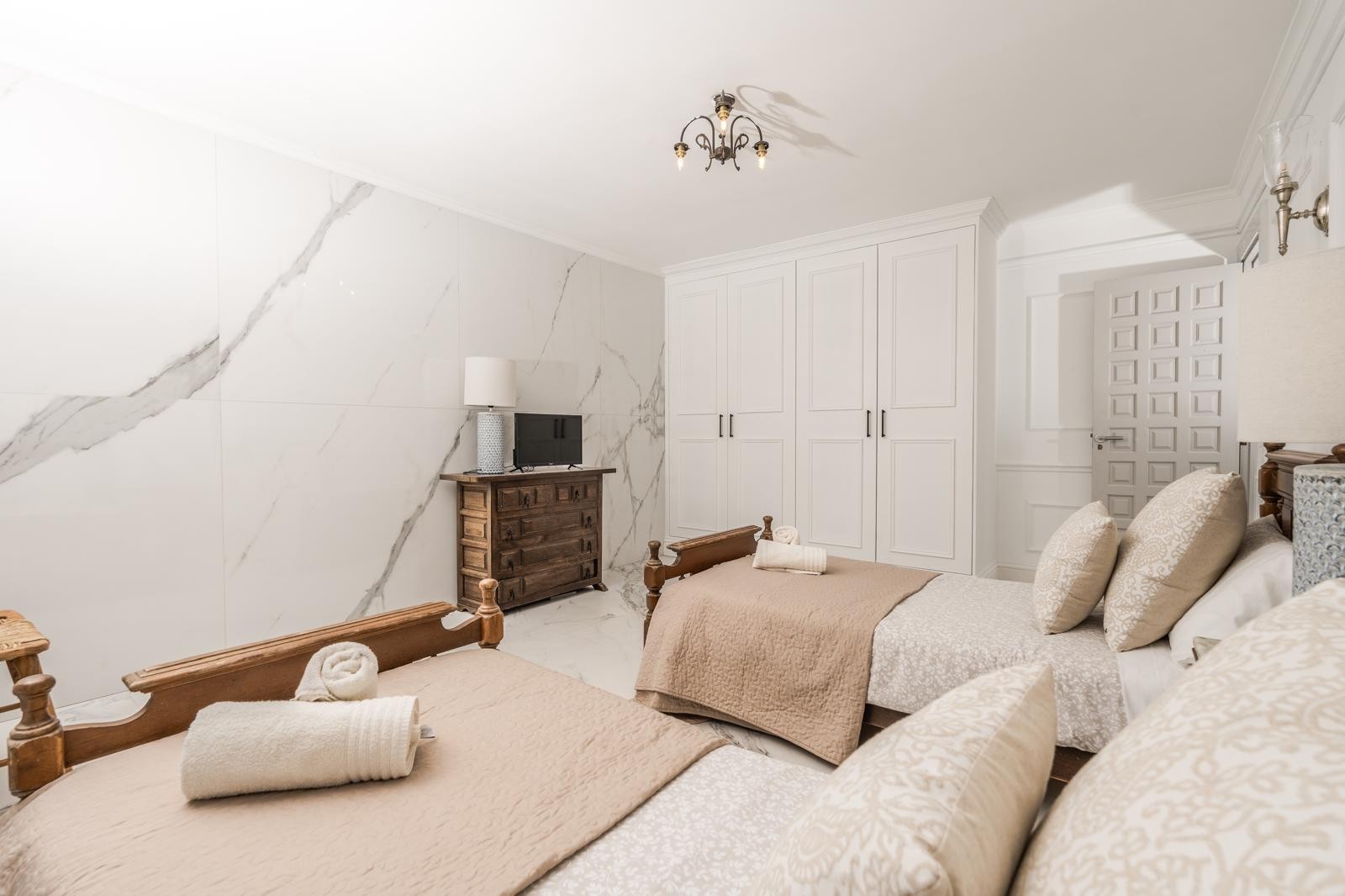Helles Schlafzimmer mit zwei Einzelbetten, einer Wandverkleidung in Marmoroptik, weißen Einbauschränken und rustikalen Holzmöbeln.