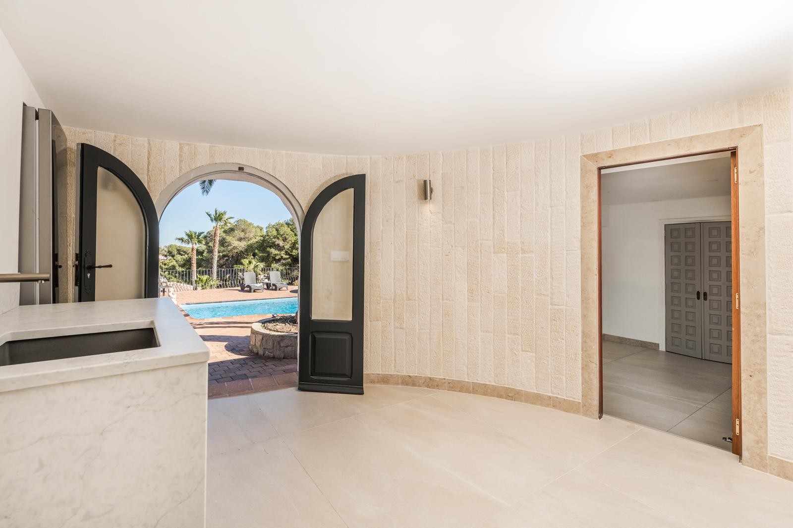 Innenbereich mit Natursteinwänden und Marmor-Arbeitsplatte, der durch einen Rundbogen direkten Zugang zur Poolterrasse und dem Garten bietet.