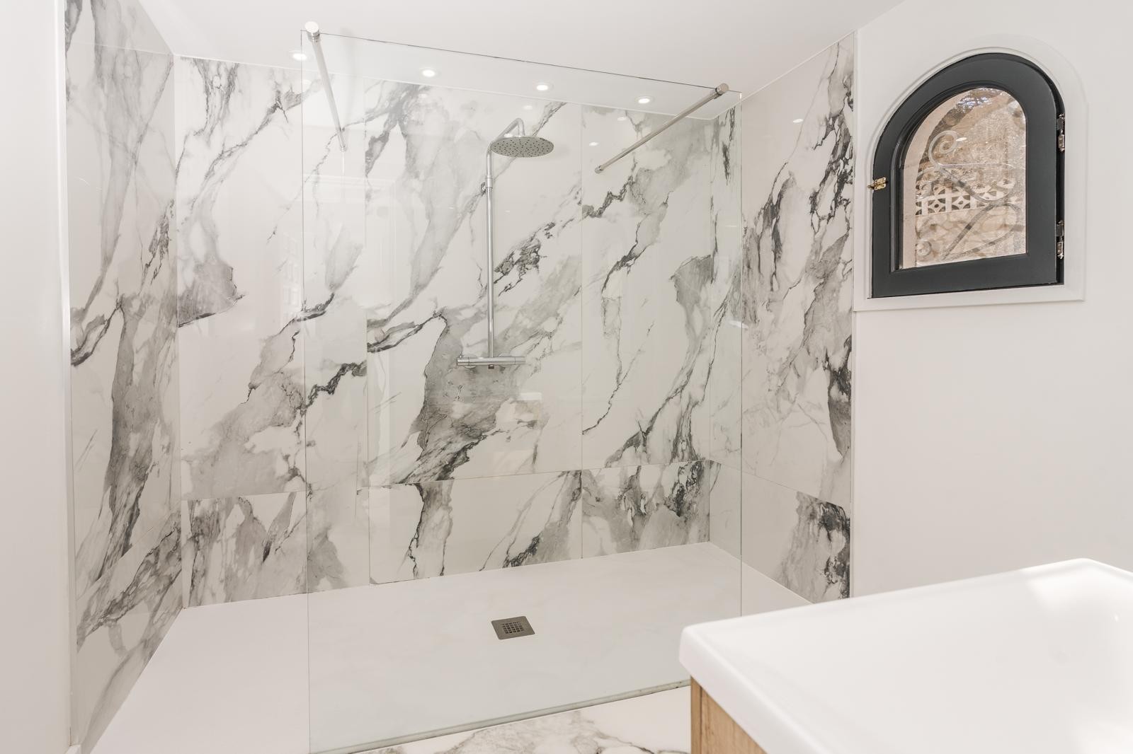 Modernes Badezimmer mit begehbarer Dusche, großformatigen Marmorfliesen, Glasabtrennung und einem Rundbogenfenster mit schwarzem Rahmen.