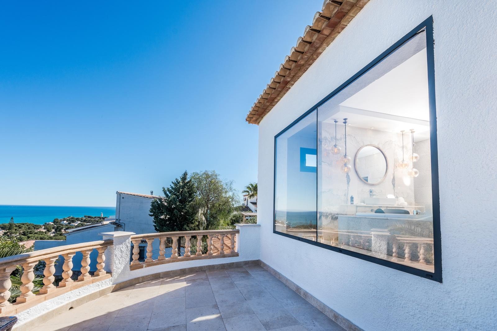 Obere Terrasse mit Steinbalustrade und Meerblick, neben einem großen Fenster, das den Blick auf ein modernes Marmorbad mit Designerleuchten freigibt.