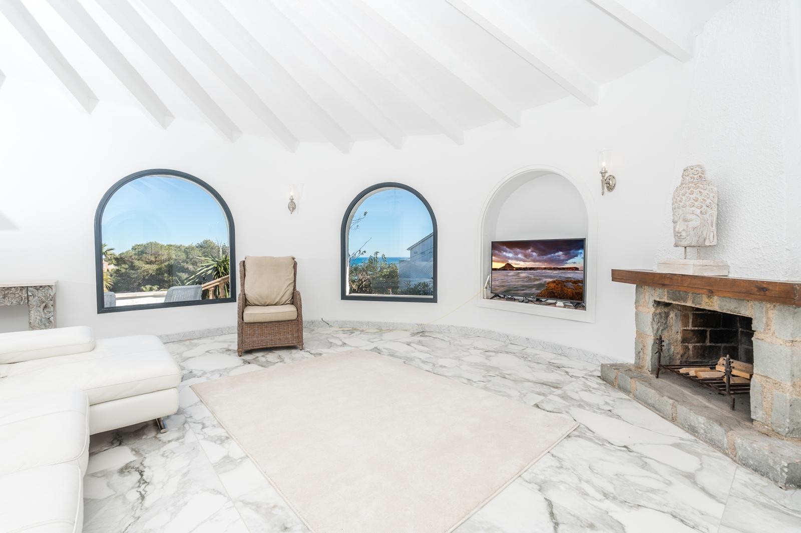 Rundes Wohnzimmer mit weißer Balkendecke, Marmorboden und Natursteinkamin. Rundbogenfenster bieten Ausblicke auf die mediterrane Umgebung und das Meer.