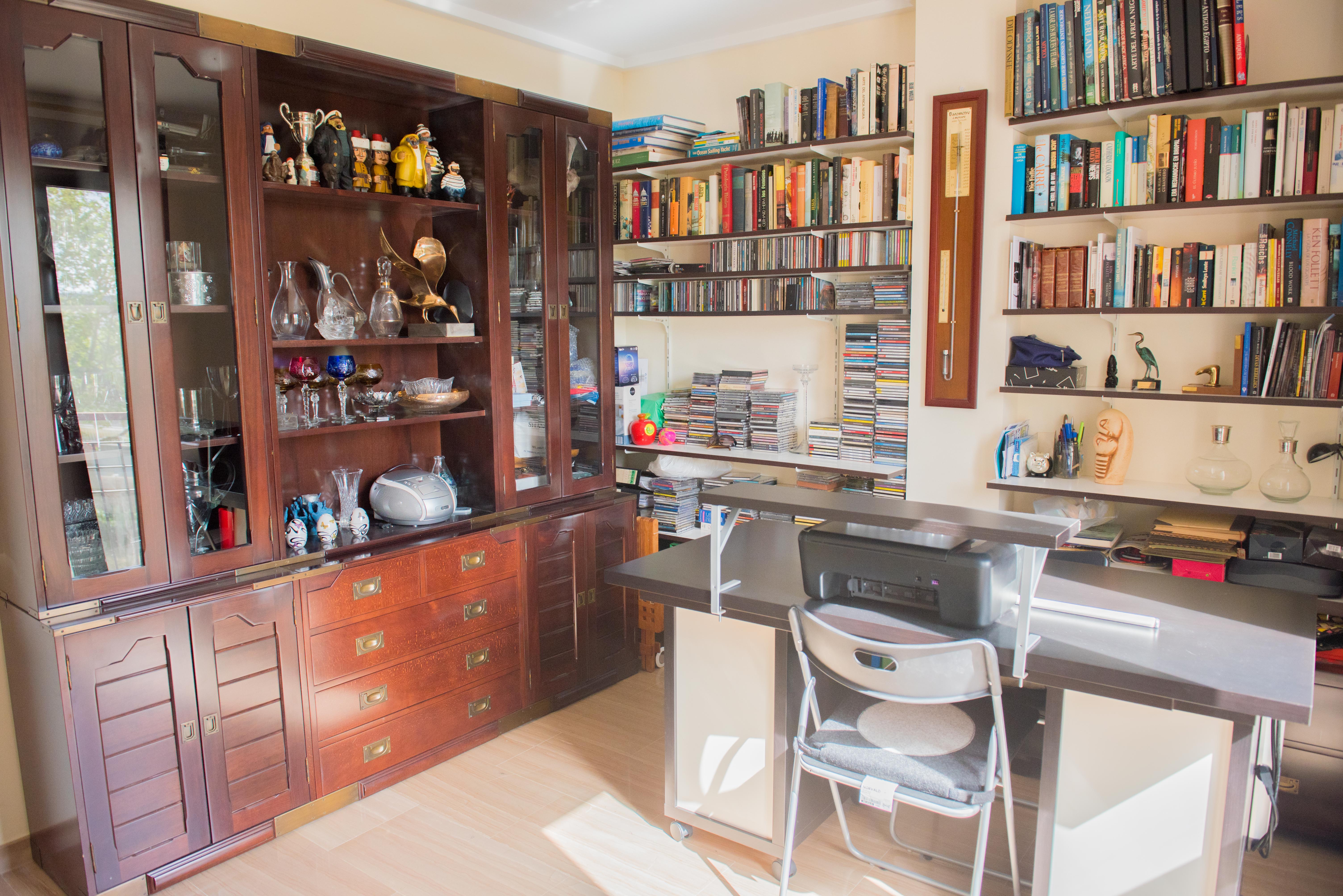 Arbeitszimmer mit einem großen Vitrinenschrank aus dunklem Holz, umfangreichen Wandregalen für Bücher und einem funktionalen Schreibtisch bei natürlichem Licht.