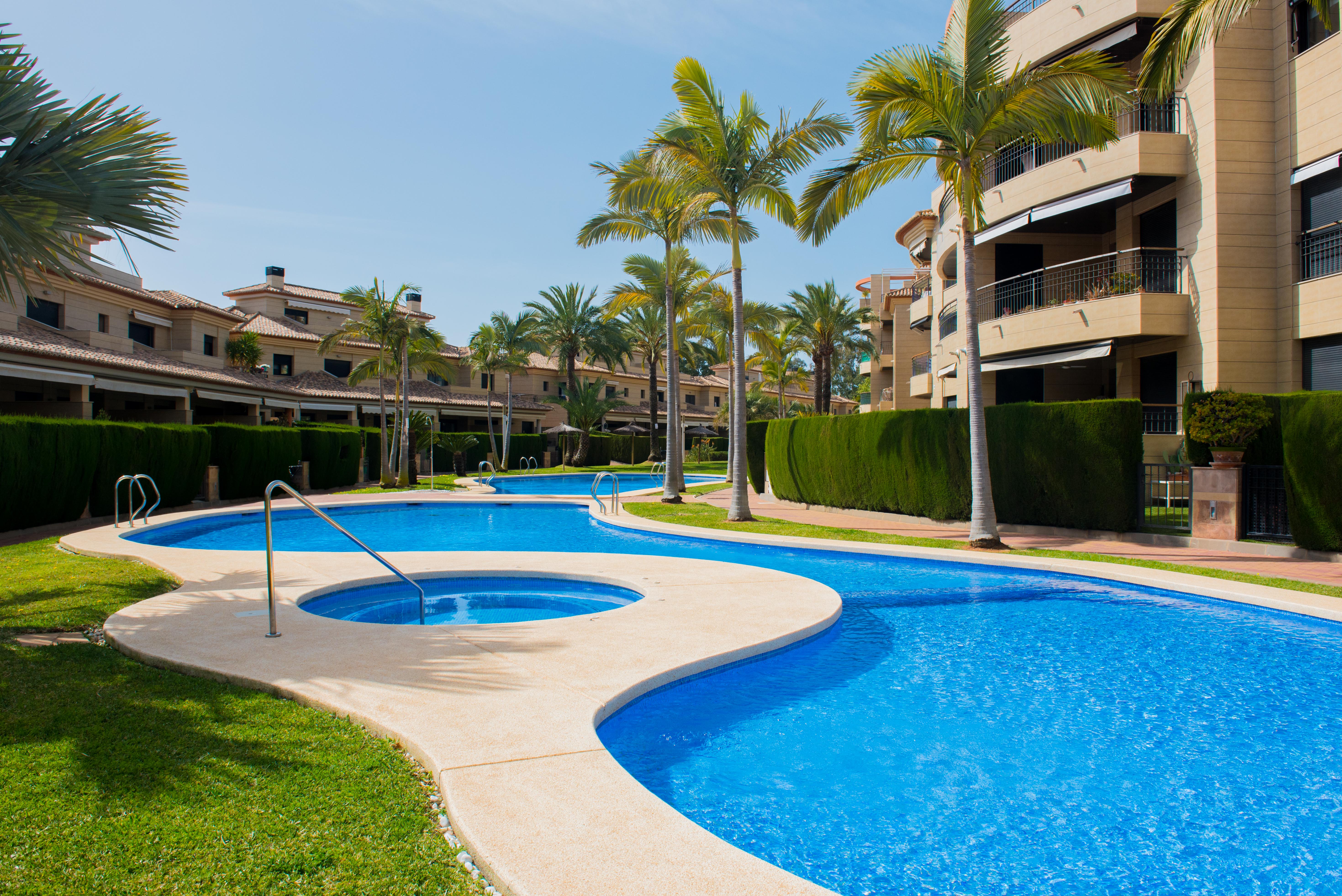 Geschwungener Gemeinschaftspool mit integriertem Whirlpool, umgeben von gepflegten Rasenflächen, Palmen und modernen Apartmenthäusern mit Steinfassaden.