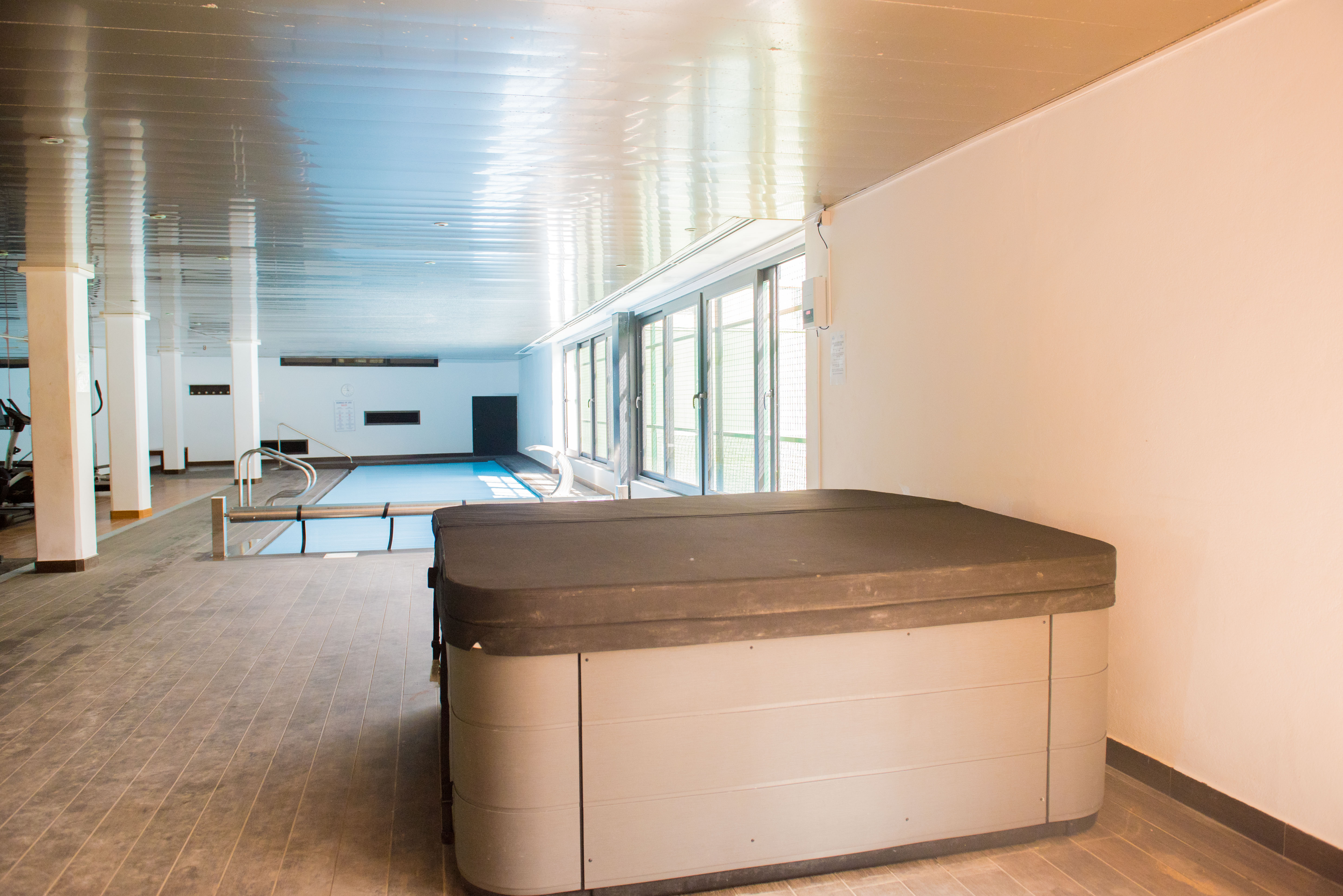 Indoor-Wellnessbereich mit abgedecktem Whirlpool, Schwimmbecken und Fitnessbereich, beleuchtet durch große Fensterfronten.