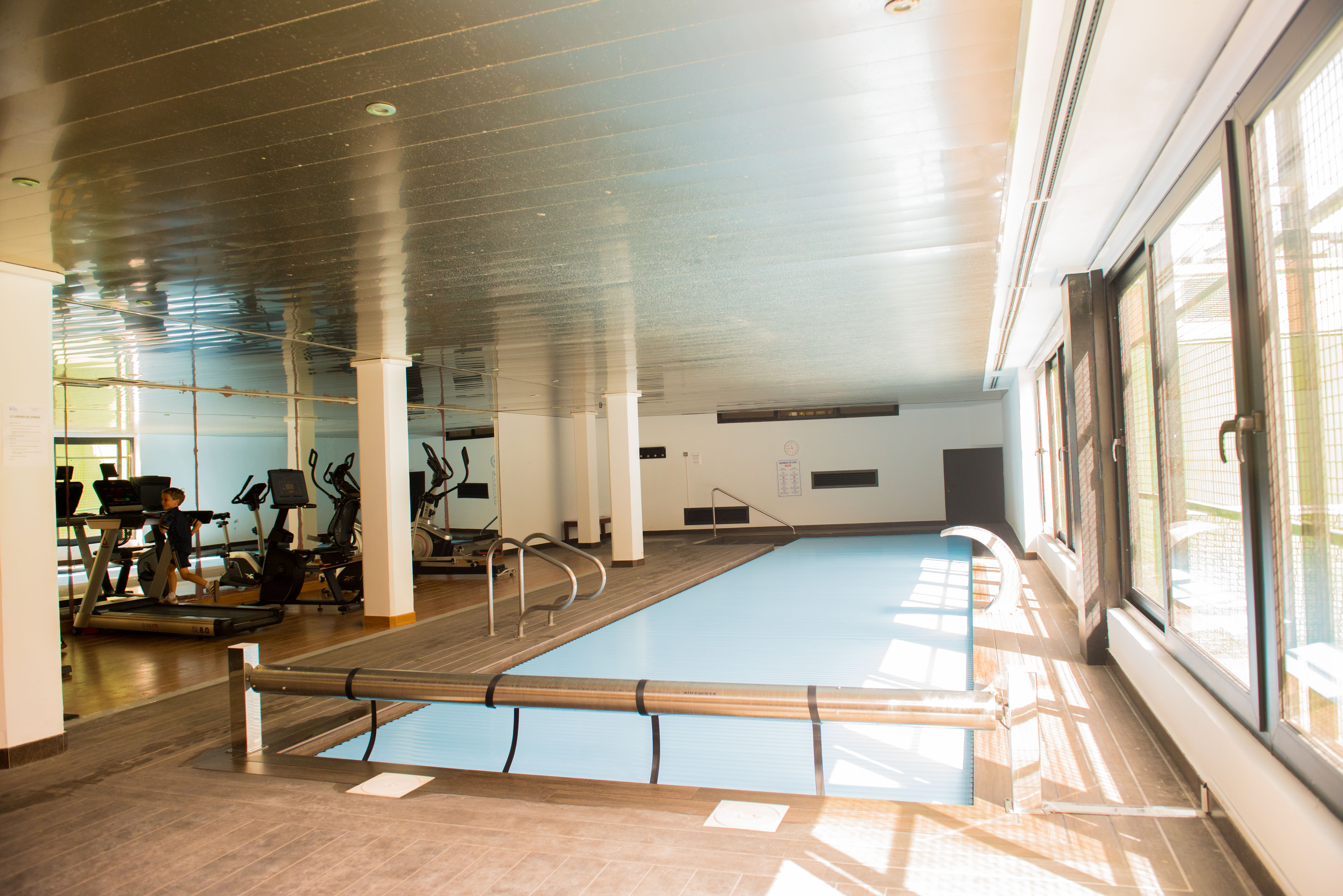 Indoor-Wellnessbereich mit beheiztem Pool und Abdeckung sowie einem Fitnessraum mit Kardiogeräten, hell beleuchtet durch große Fensterflächen.