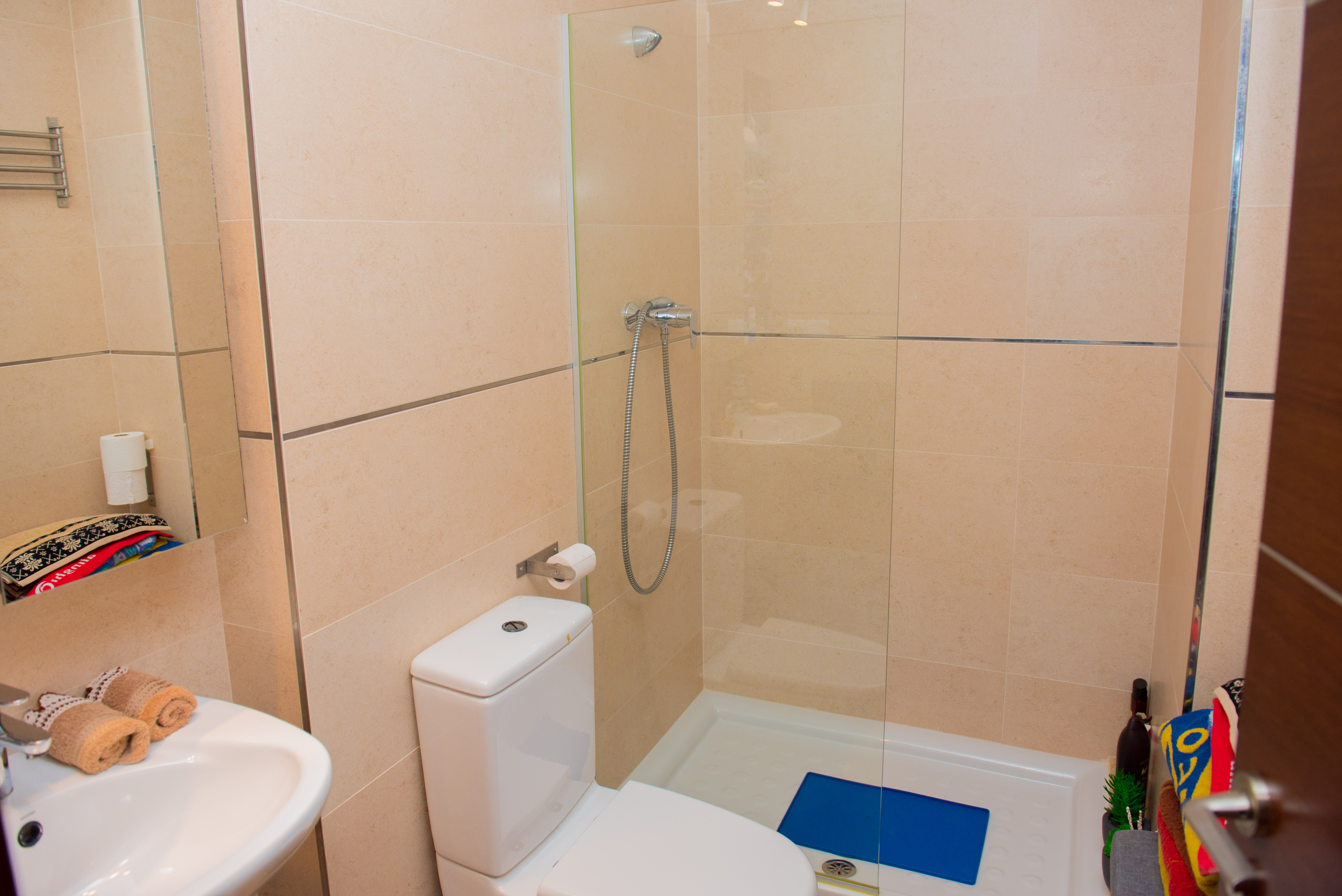 Modernes Badezimmer mit begehbarer Dusche, Glastrennwand, weißer Toilette und cremefarbenen Wandfliesen mit dekorativen Chromleisten.