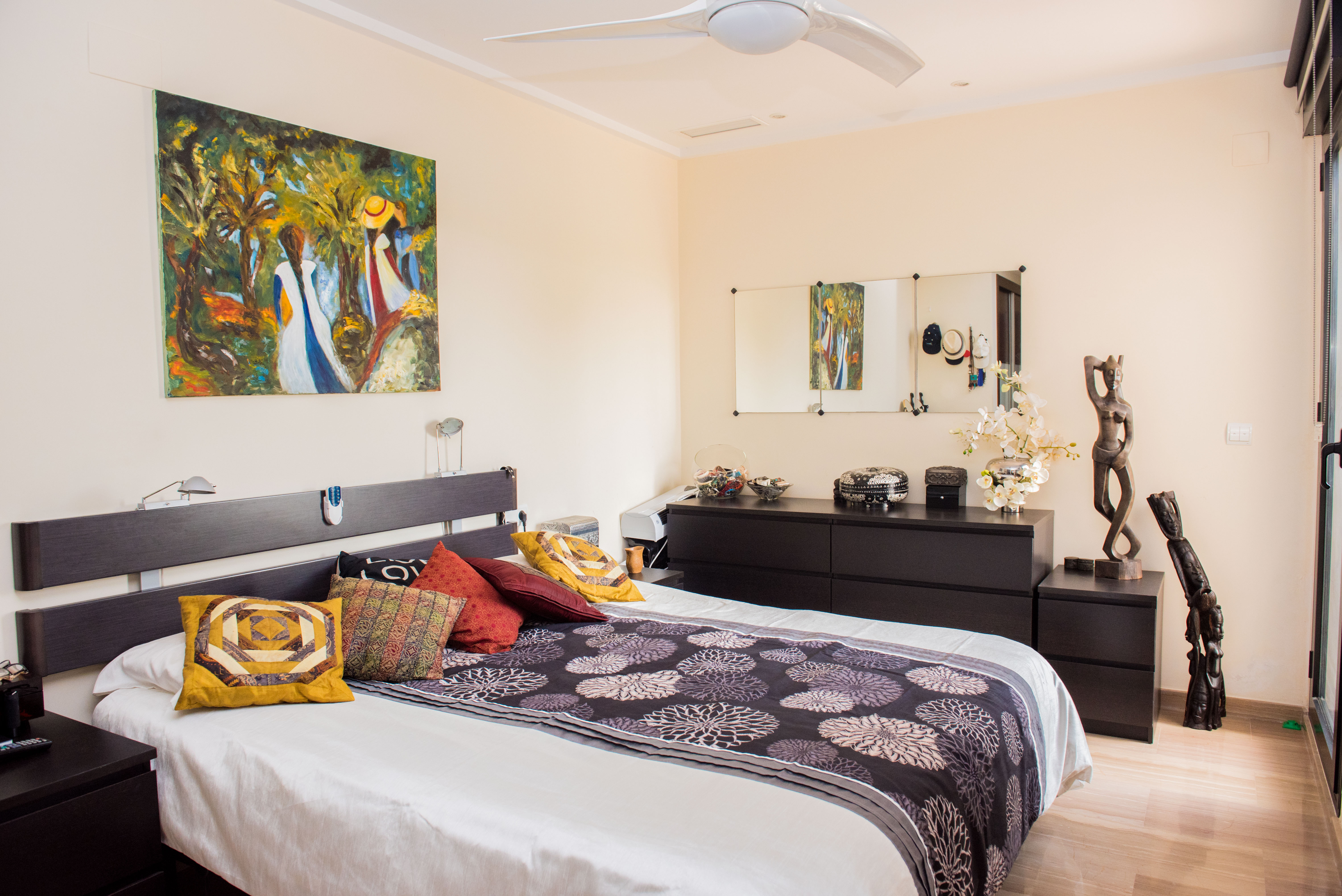 Schlafzimmer mit dunklem Holzkopfteil, passender Kommode und Deckenventilator. Ausgestattet mit farbenfroher Wandkunst und hellem Bodenbelag in Holzoptik.