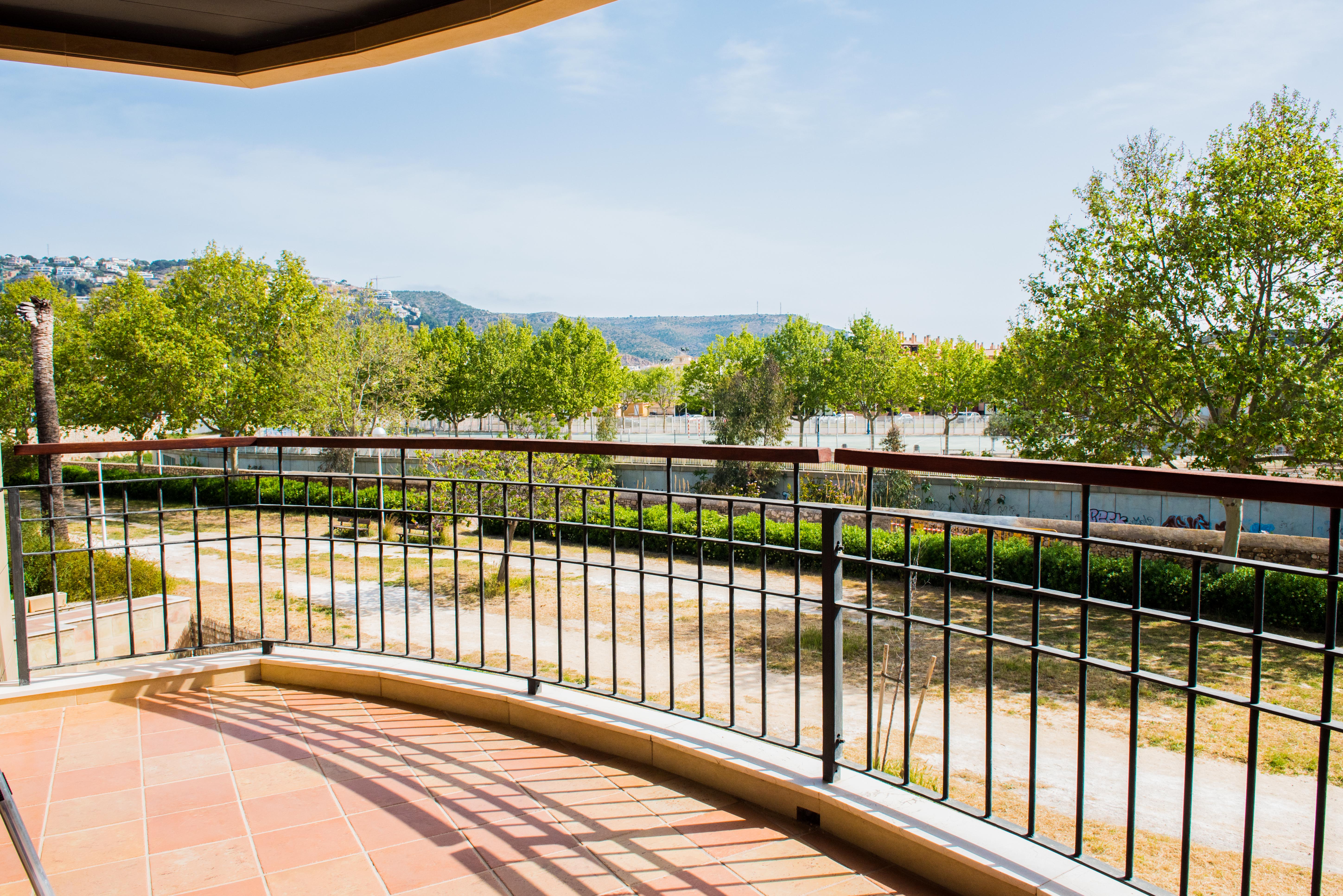 Überdachte Terrasse mit Terrakotta-Fliesenboden und Schmiedeeisengeländer mit Holzhandlauf, mit Blick auf eine Parkanlage und die Berge von Jávea.