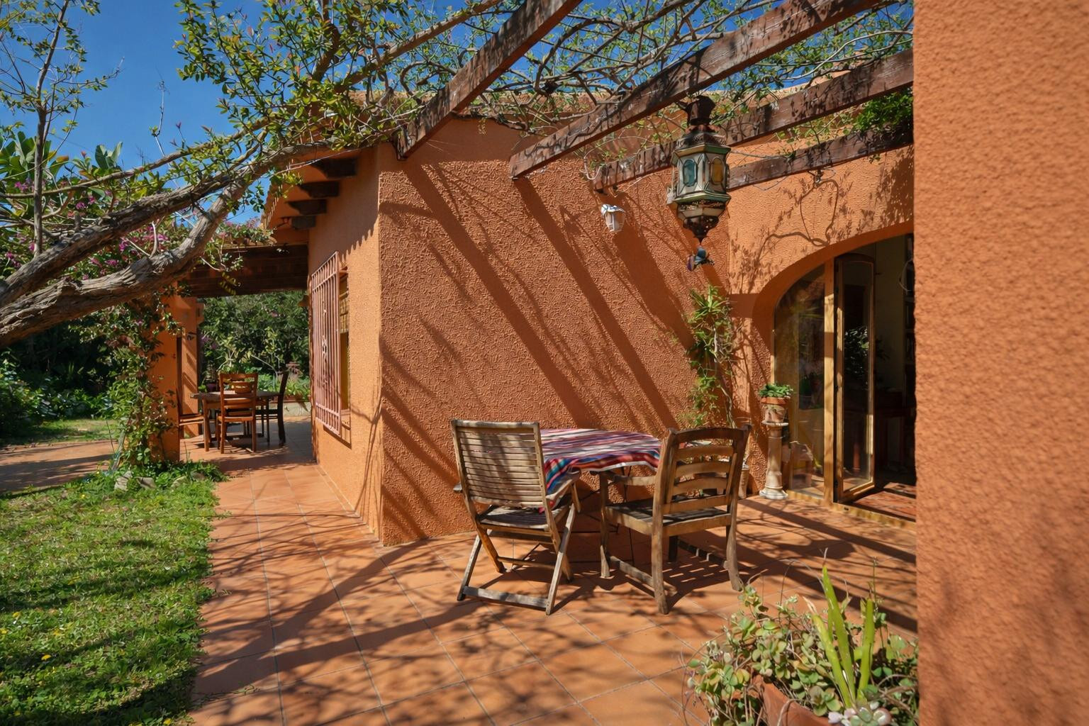 Außen-Terrasse mit Terrakotta-Fliesen, einer Holzpergola mit Kletterpflanzen und rustikalen Holzmöbeln vor einer warmfarbenen Fassade.