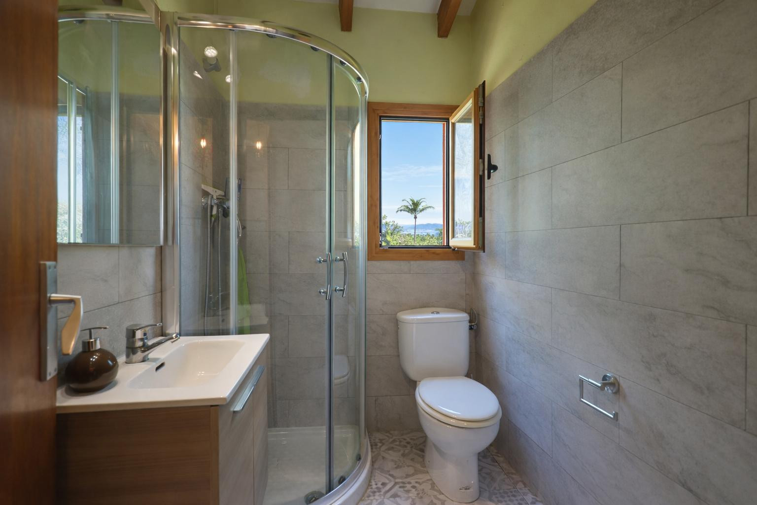 Funktionales Badezimmer mit einer runden Glasduschkabine, grauen Fliesen und Holzbalken. Das Fenster bietet Tageslicht und einen Blick auf die mediterrane Umgebung.