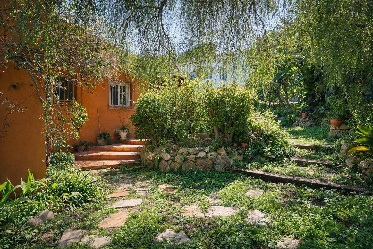 Mediterraner Garten mit Steinpfaden und Holzstufen, umgeben von üppiger Vegetation und Natursteinmauern vor der terracottafarbenen Fassade.