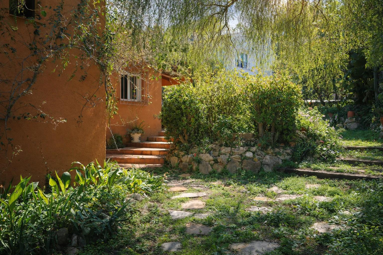 Naturbelassener Garten mit Steinpfaden und Terrakottastufen, die zur ockerfarbenen Fassade der Villa führen, umgeben von üppiger mediterraner Vegetation.