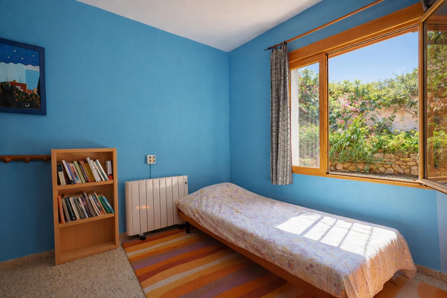 Schlafzimmer mit leuchtend blauen Wänden, Einzelbett und Holzregal. Das Holzfenster bietet einen Blick auf den Garten mit seinen charakteristischen Natursteinmauern.