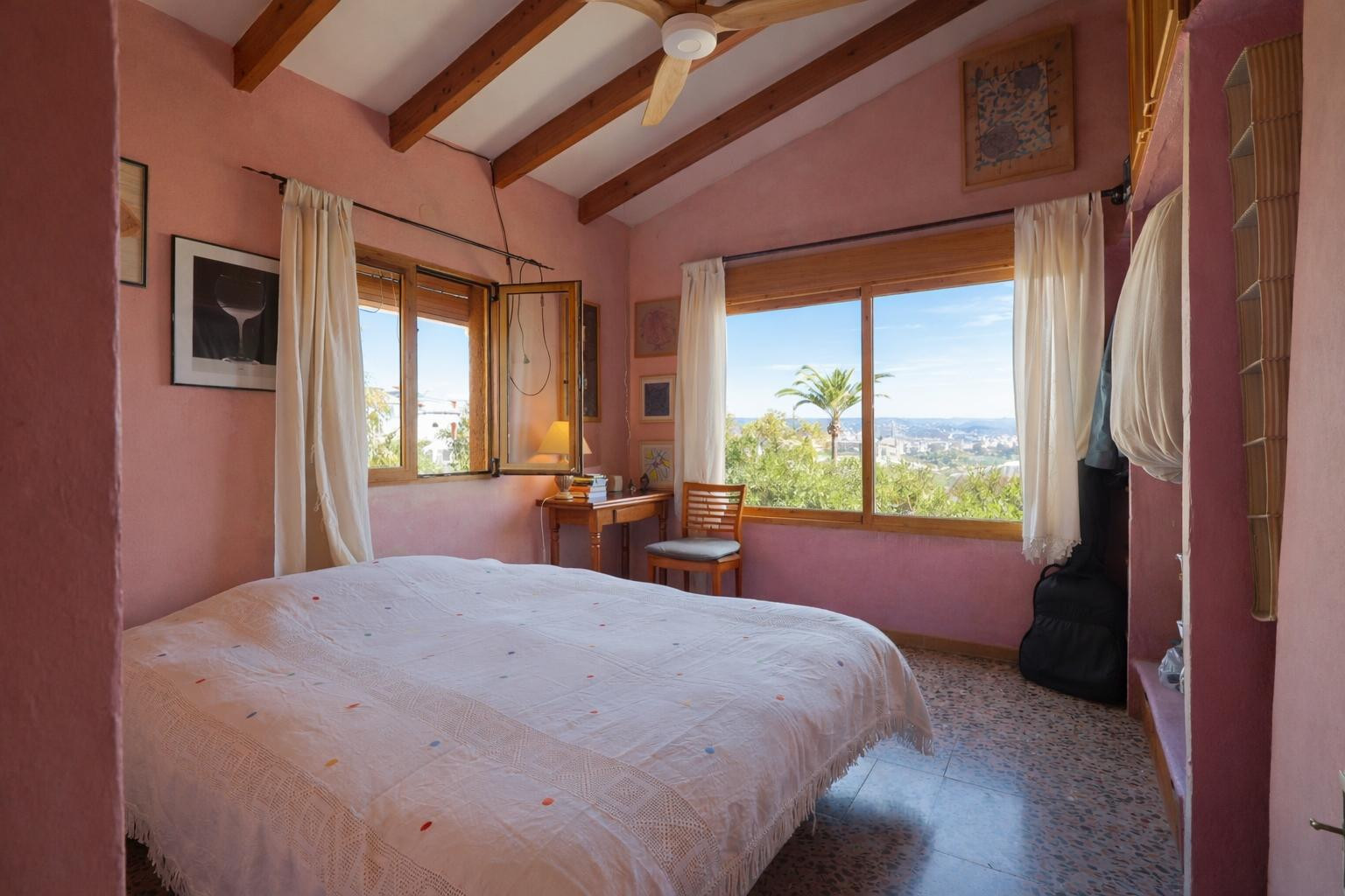 Schlafzimmer mit Sichtbalkendecke, warmen Wandfarben und großen Fenstern mit weitem Blick auf die mediterrane Landschaft von Jávea.
