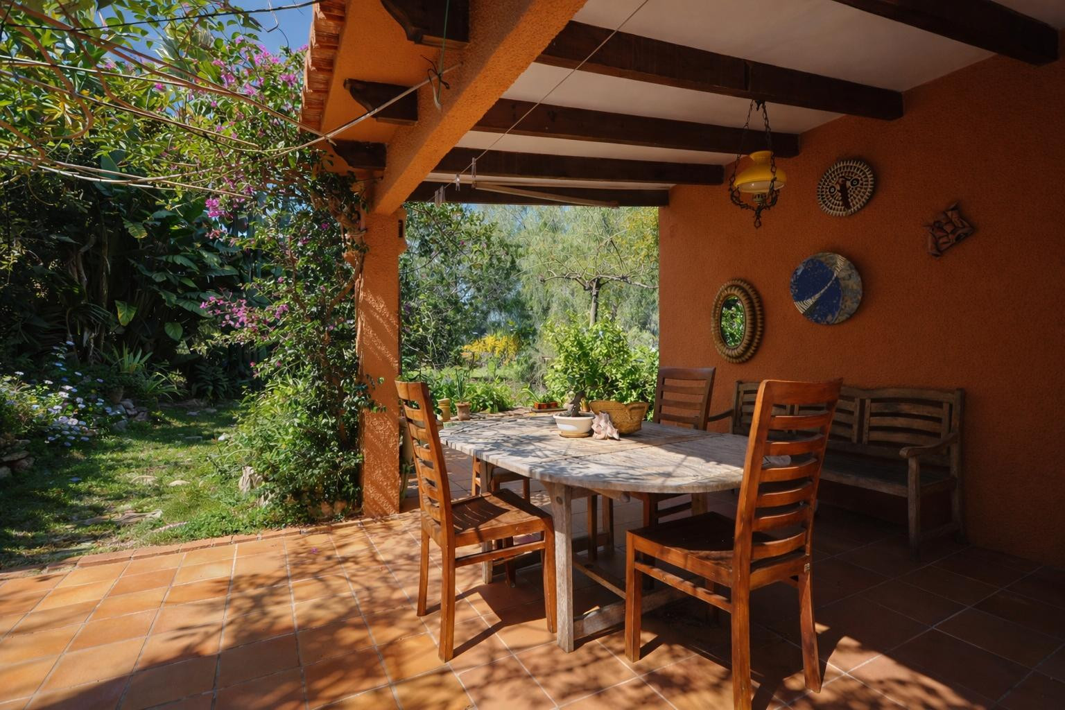 Überdachte Terrasse mit Holzbalkendecke, Terrakottaboden und Essbereich im Freien, umgeben von einem üppigen mediterranen Garten.