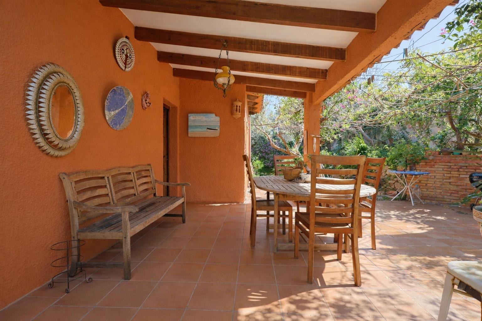 Überdachte Terrasse mit sichtbaren Holzbalken, Terrakottaboden und Essbereich im Freien vor warmen, ockerfarbenen Wänden.