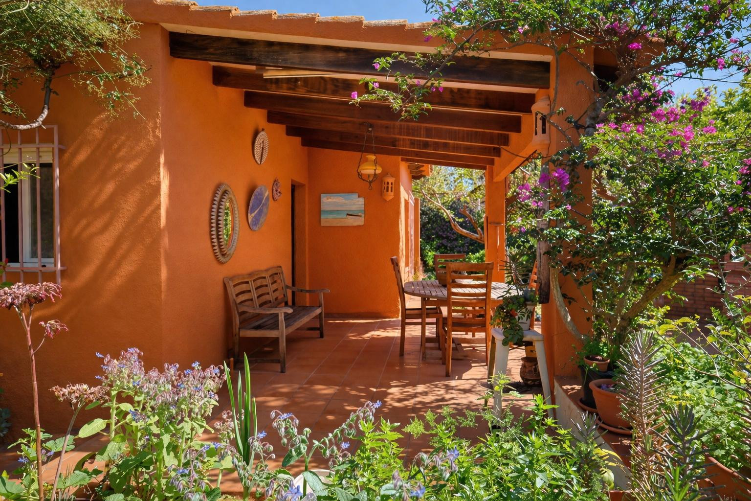 Überdachte Terrasse mit sichtbaren Holzbalken, Terrakottaboden und Holzmöbeln, eingebettet in einen üppigen mediterranen Garten mit Bougainvillea.