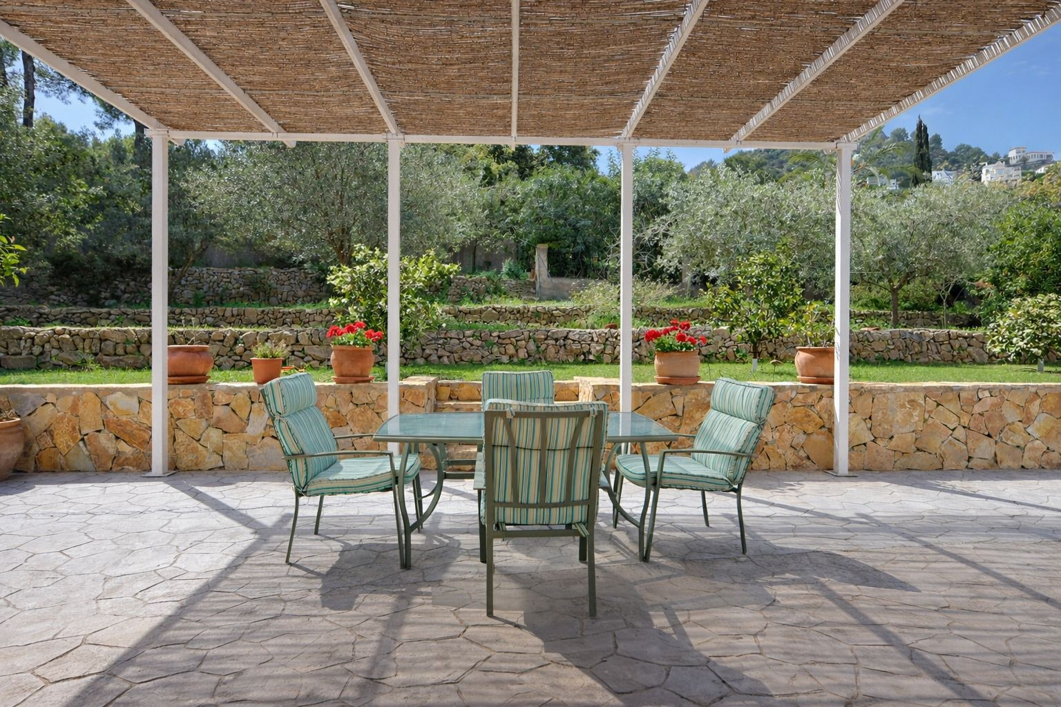 Außen-Terrasse mit Schilfrohr-Pergola, Essbereich und Blick auf einen terrassierten Garten mit Natursteinmauern und Obstbäumen.