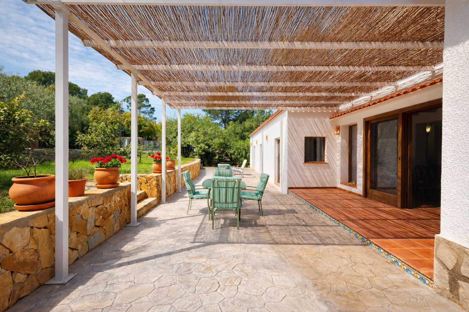 Außen-Terrasse mit Schilfrohr-Pergola, Steinboden und Gartenmöbeln. Eine Natursteinmauer mit Terrakotta-Töpfen grenzt an den gepflegten Garten an.