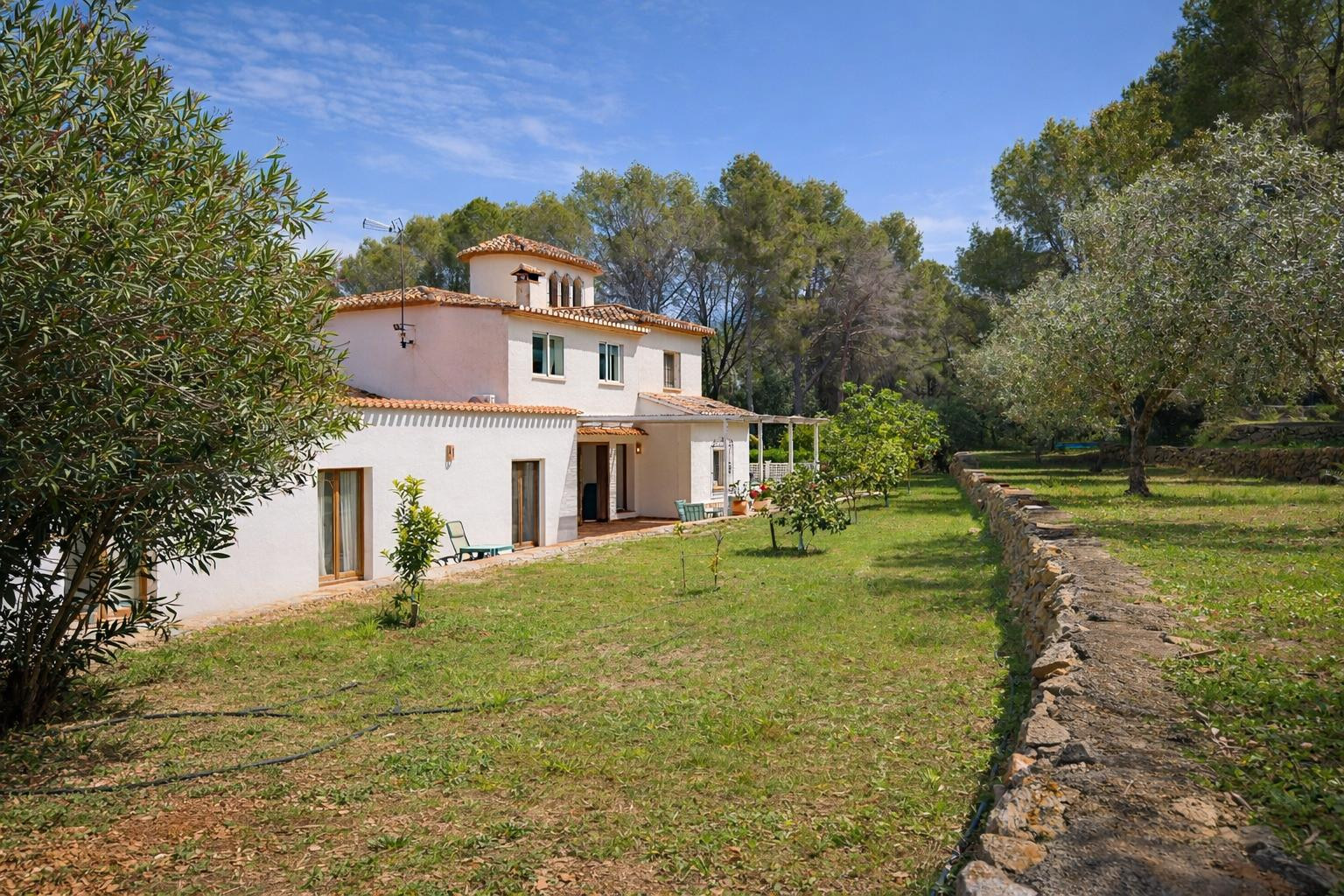 Außenansicht der weißen Villa mit Terrakotta-Ziegeldach, eingebettet in einen Garten mit Obstbäumen und einer charakteristischen Natursteinmauer.