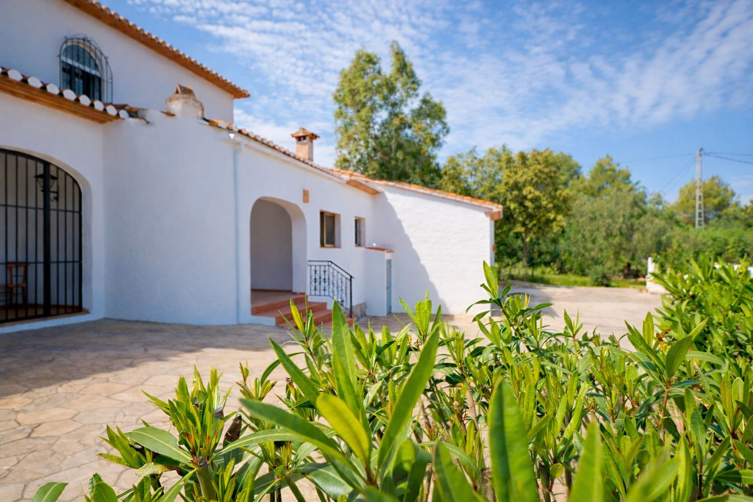 Außenansicht der weißen Villa mit traditionellem Ziegeldach, Natursteinpflaster und Rundbogen-Eingängen, eingebettet in eine grüne Gartenlandschaft.