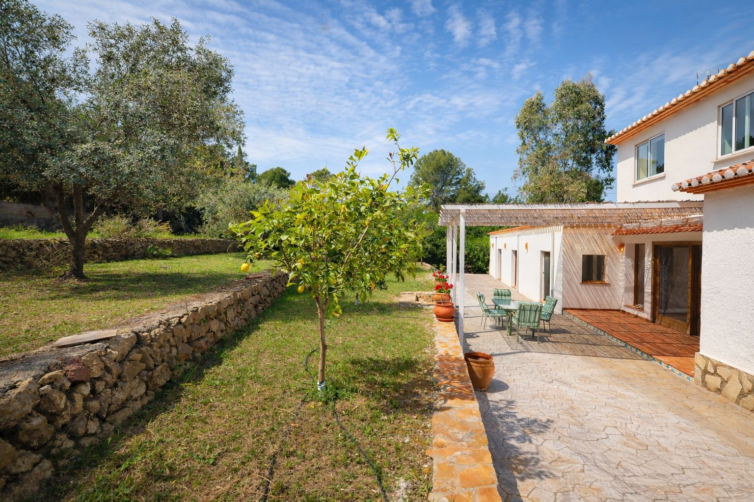 Außenansicht mit gepflasterter Terrasse, schattiger Pergola und terrassiertem Garten mit Natursteinmauern und Obstbäumen.