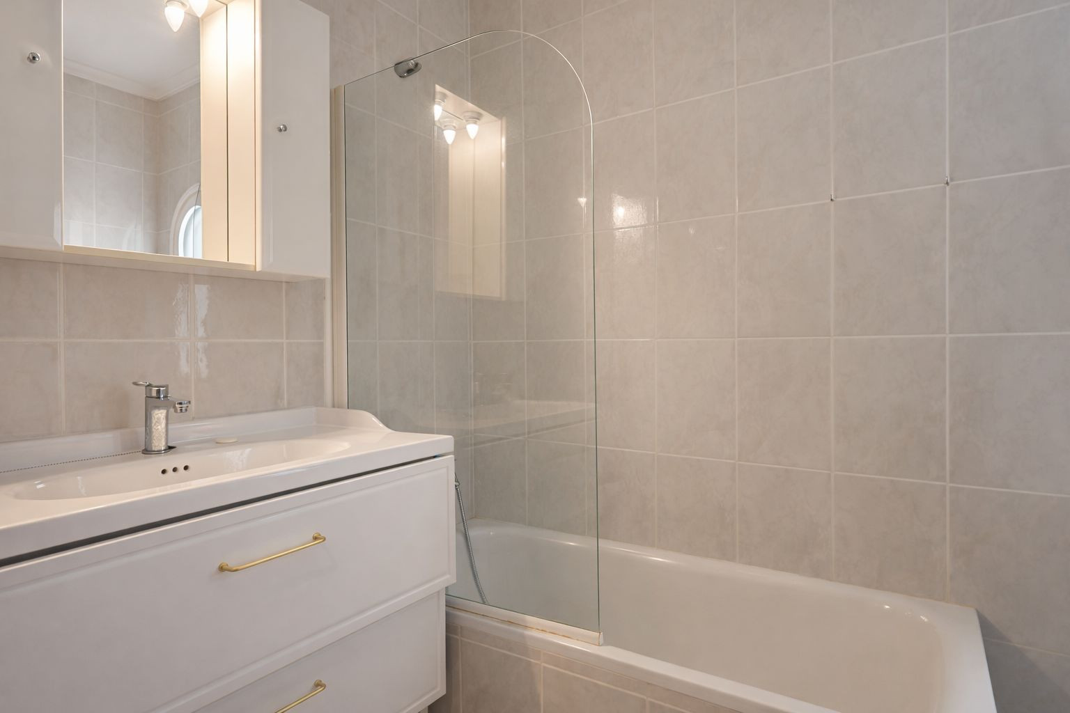 Badezimmer mit Badewanne und gebogener Glaswand, weißem Unterschrank mit goldenen Griffen und einem Spiegelschrank mit integrierter Beleuchtung.