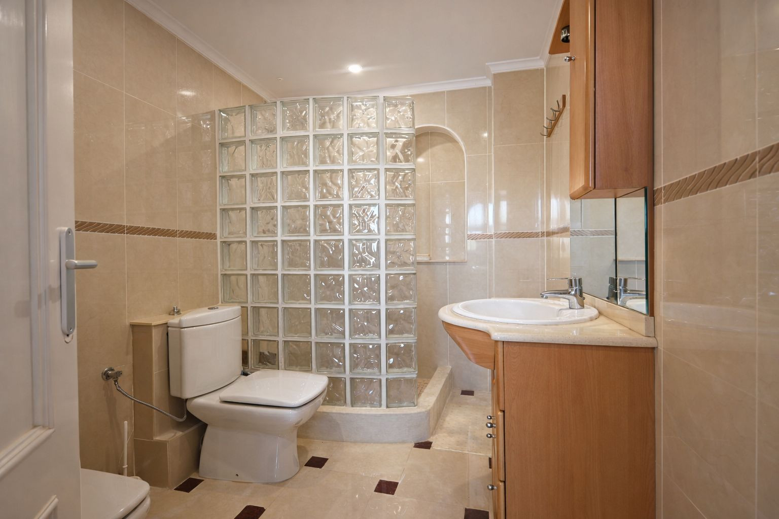 Badezimmer mit cremefarbenen Wandfliesen, einer Dusche mit Glasbausteinwand und einem Waschtisch aus Holz mit integriertem Spiegel.