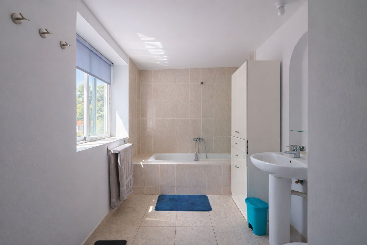 Funktionales Badezimmer mit Einbaubadewanne, neutralen Keramikfliesen, Standwaschbecken und Fenster für natürliches Tageslicht.