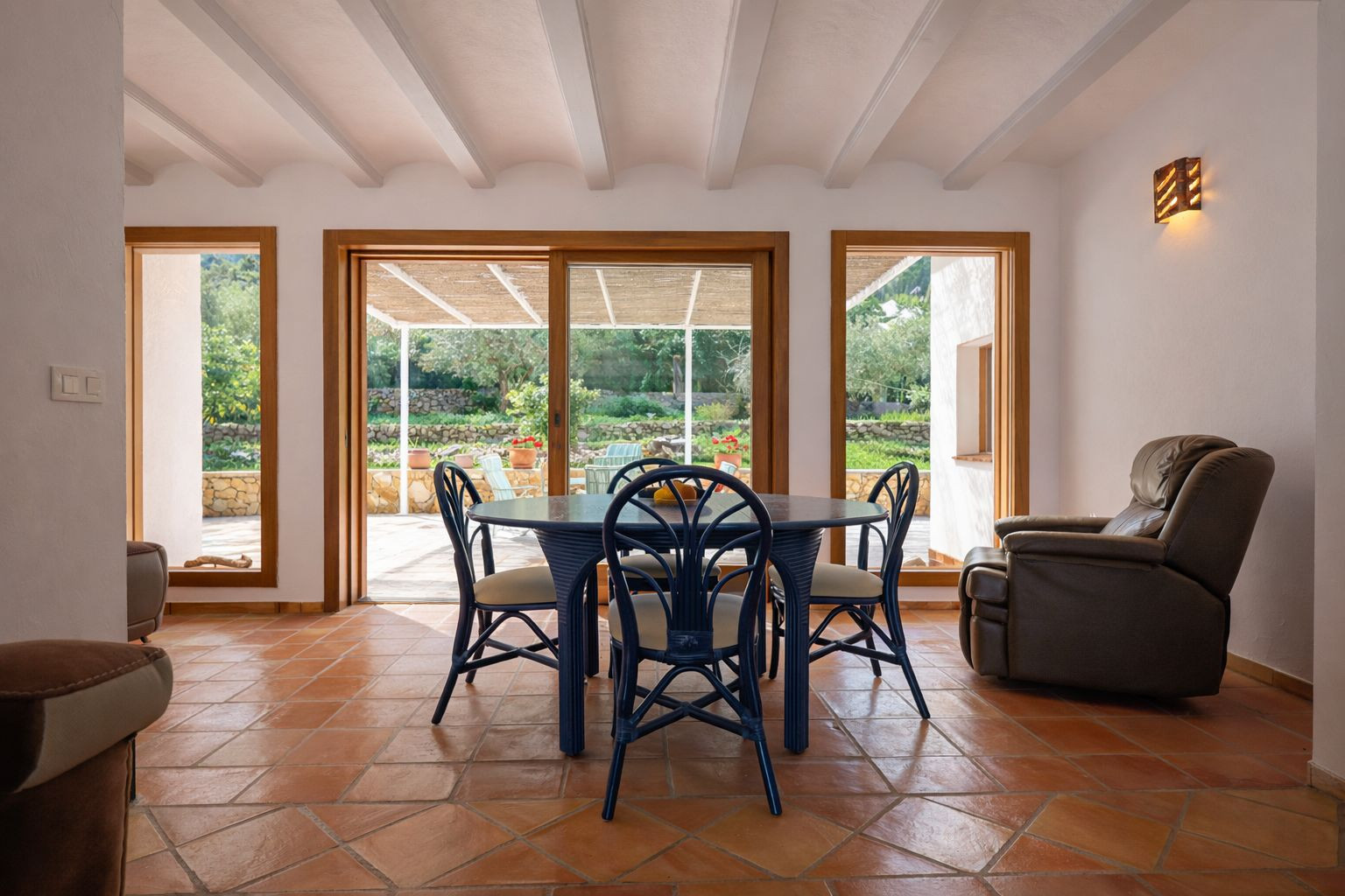 Heller Essbereich mit weißen Sichtbalken, Terrakotta-Fliesenboden und direktem Zugang zur überdachten Terrasse durch große Holzschiebefenster.