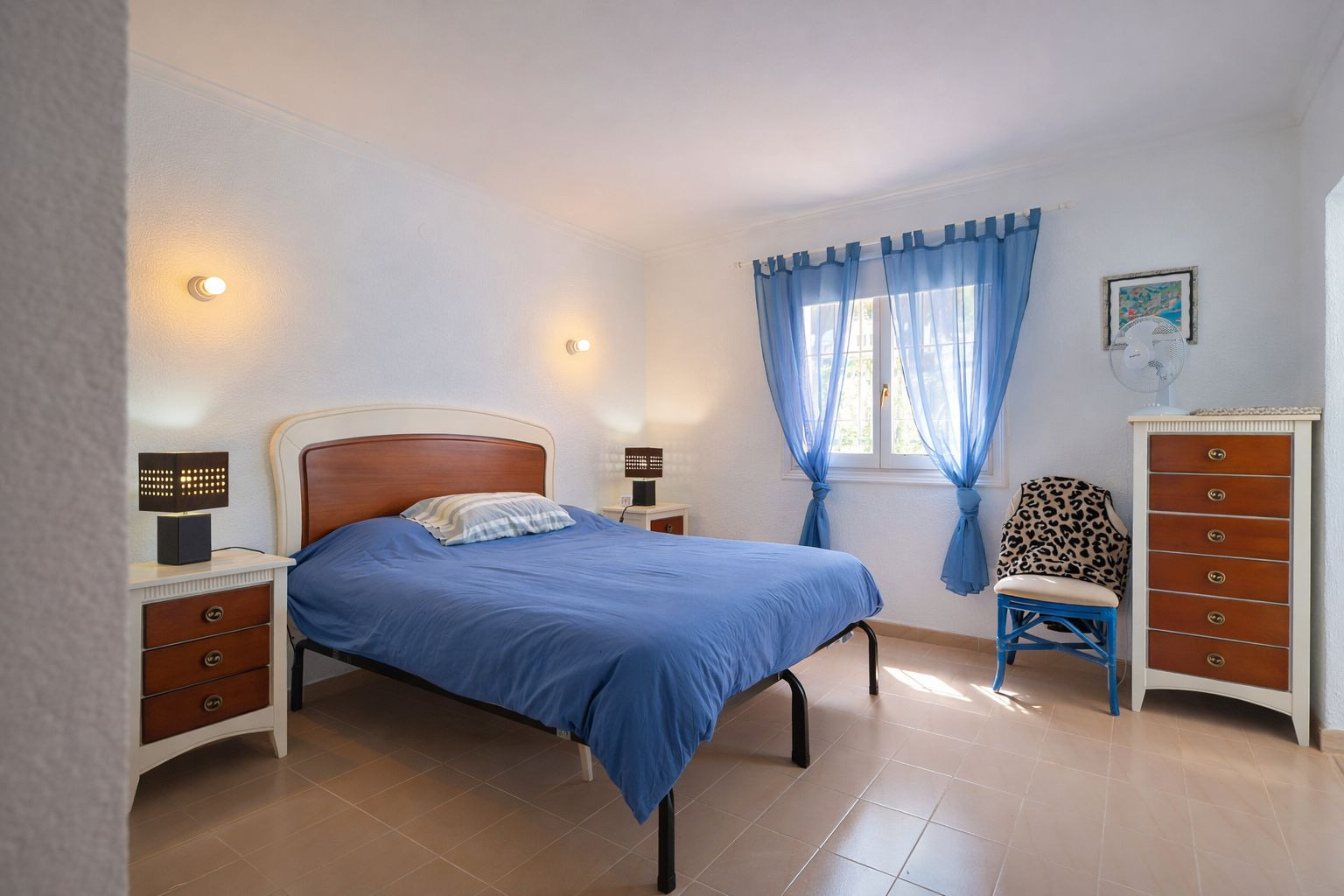 Helles Schlafzimmer mit Fliesenboden, Doppelbett mit Holz-Kopfteil und passenden Möbeln. Ausgestattet mit einer hohen Kommode und Tageslicht durch blaue Vorhänge.