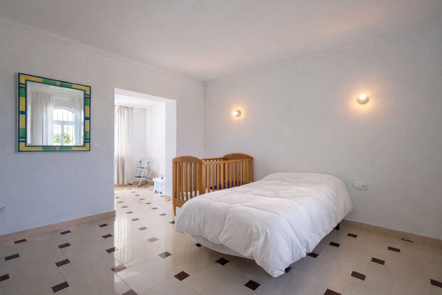 Helles Schlafzimmer mit verzierten Fliesenböden, Einzelbett und Kinderbett aus Holz. Inklusive Wandleuchten und Durchgang zu einem lichtdurchfluteten Nebenraum.