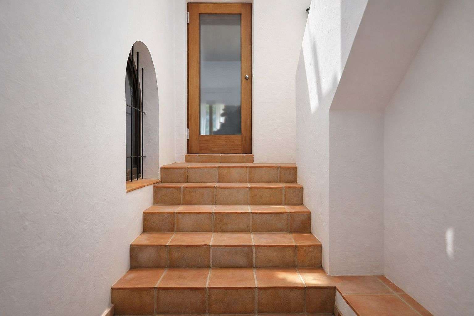 Innentreppe mit Stufen aus Terrakotta, die zu einer Holztür mit Glaseinsatz führt, ergänzt durch ein Rundbogenfenster mit schmiedeeisernem Gitter.