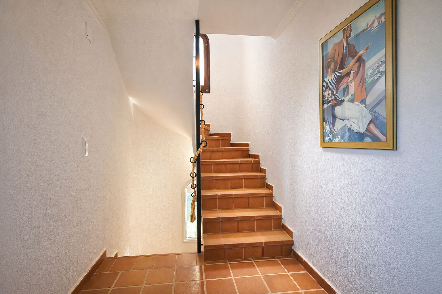 Treppe mit Terrakotta-Fliesen, schwarzem Metallgeländer mit Seildetails und strukturierten weißen Wänden, ergänzt durch natürliches Licht.