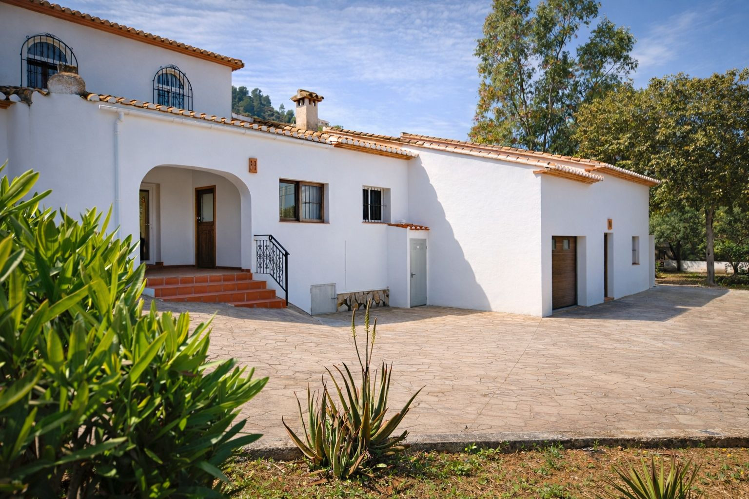 Weiße Fassade im mediterranen Stil mit Terrakotta-Ziegeldach, bogenförmigem Eingangsbereich und großzügig gepflasterter Einfahrt mit integrierter Garage.