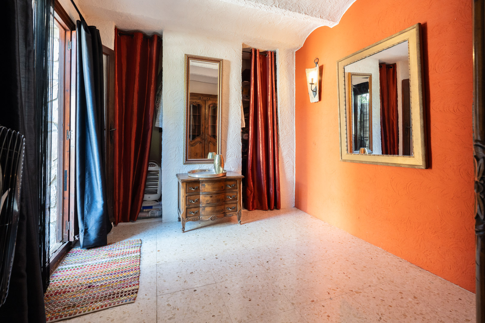 Ankleidebereich mit orangefarbener Akzentwand, verzierten Spiegeln, klassischer Holzkommode und Terrazzoboden. Rote Vorhänge verbergen die integrierten Schranknischen.