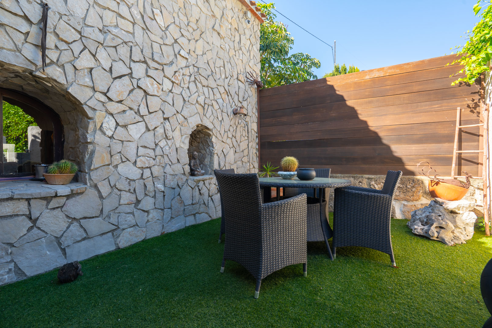 Außenbereich mit einer Natursteinmauer samt dekorativen Nischen, einer dunklen Rattan-Sitzgruppe und einem Bodenbelag aus Kunstrasen.