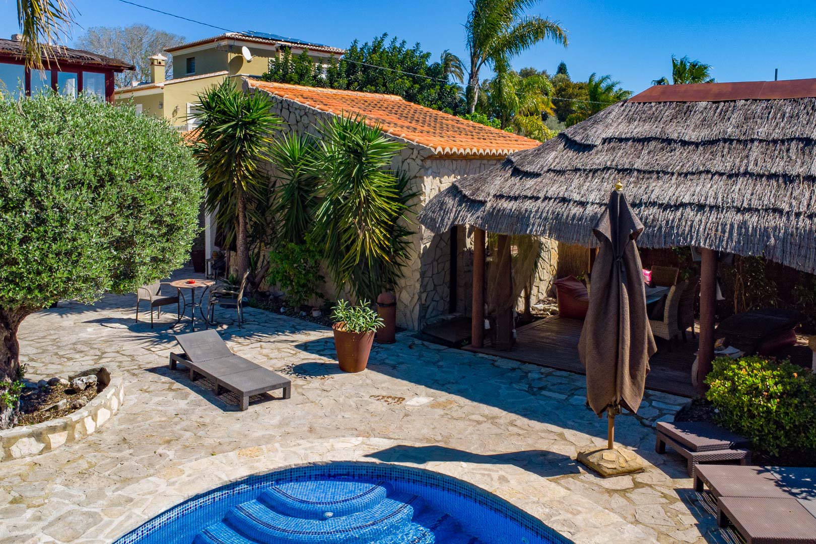 Außenbereich mit Naturstein-Terrasse, Pool und balinesischer Reetdach-Pergola neben einem rustikalen Natursteingebäude und mediterraner Bepflanzung.