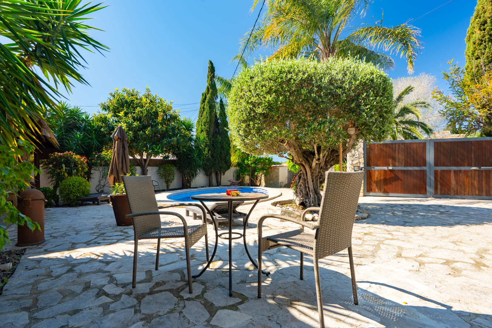 Außenbereich mit Natursteinpflaster, Rattan-Sitzgruppe und Blick auf den privaten Pool, umgeben von mediterraner Bepflanzung und einem markanten Olivenbaum.
