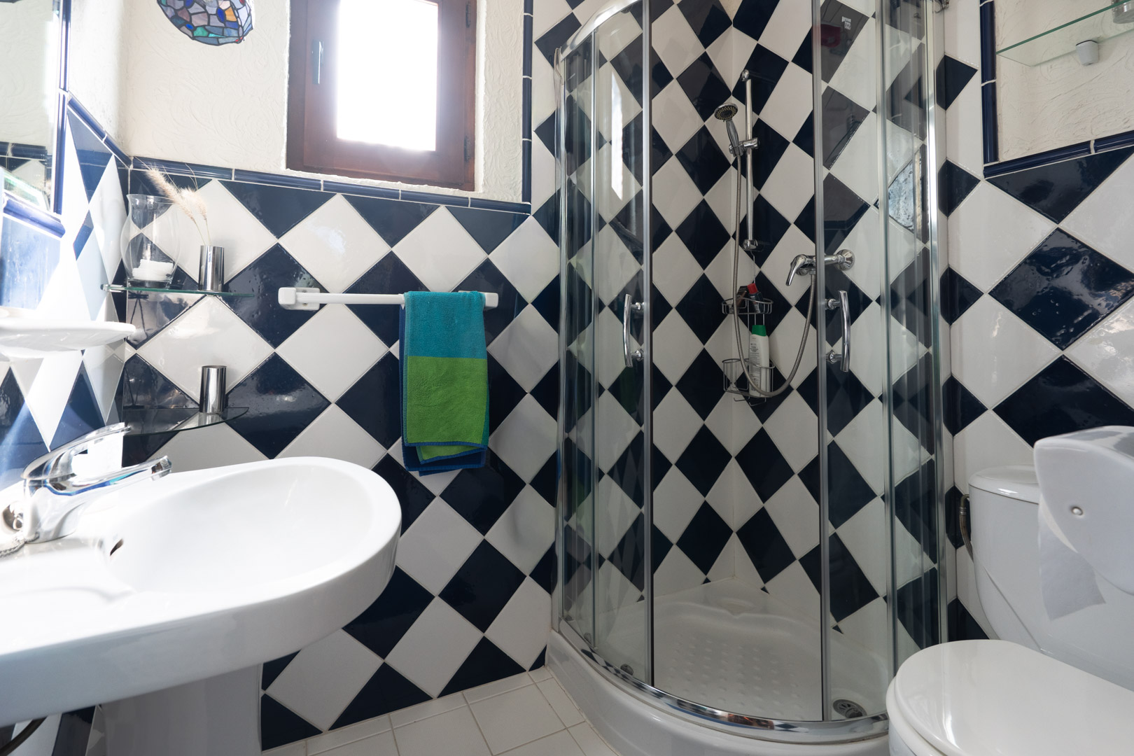 Badezimmer mit blau-weißem Karomuster an den Wänden, einer abgerundeten Eckduschkabine aus Glas und einem klassischen weißen Standwaschbecken.
