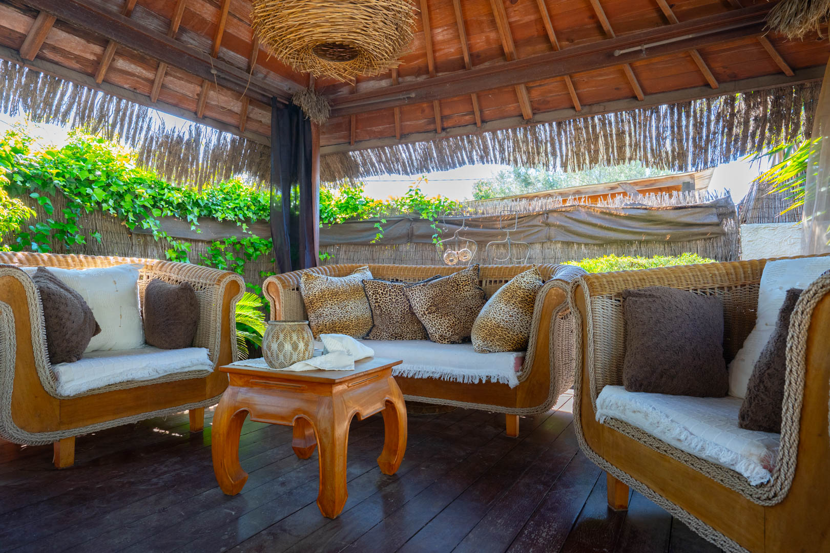 Balinesische Pergola mit sichtbarer Holzkonstruktion, Rattansofas mit dekorativen Kissen und einem Couchtisch auf einem dunklen Holzboden.