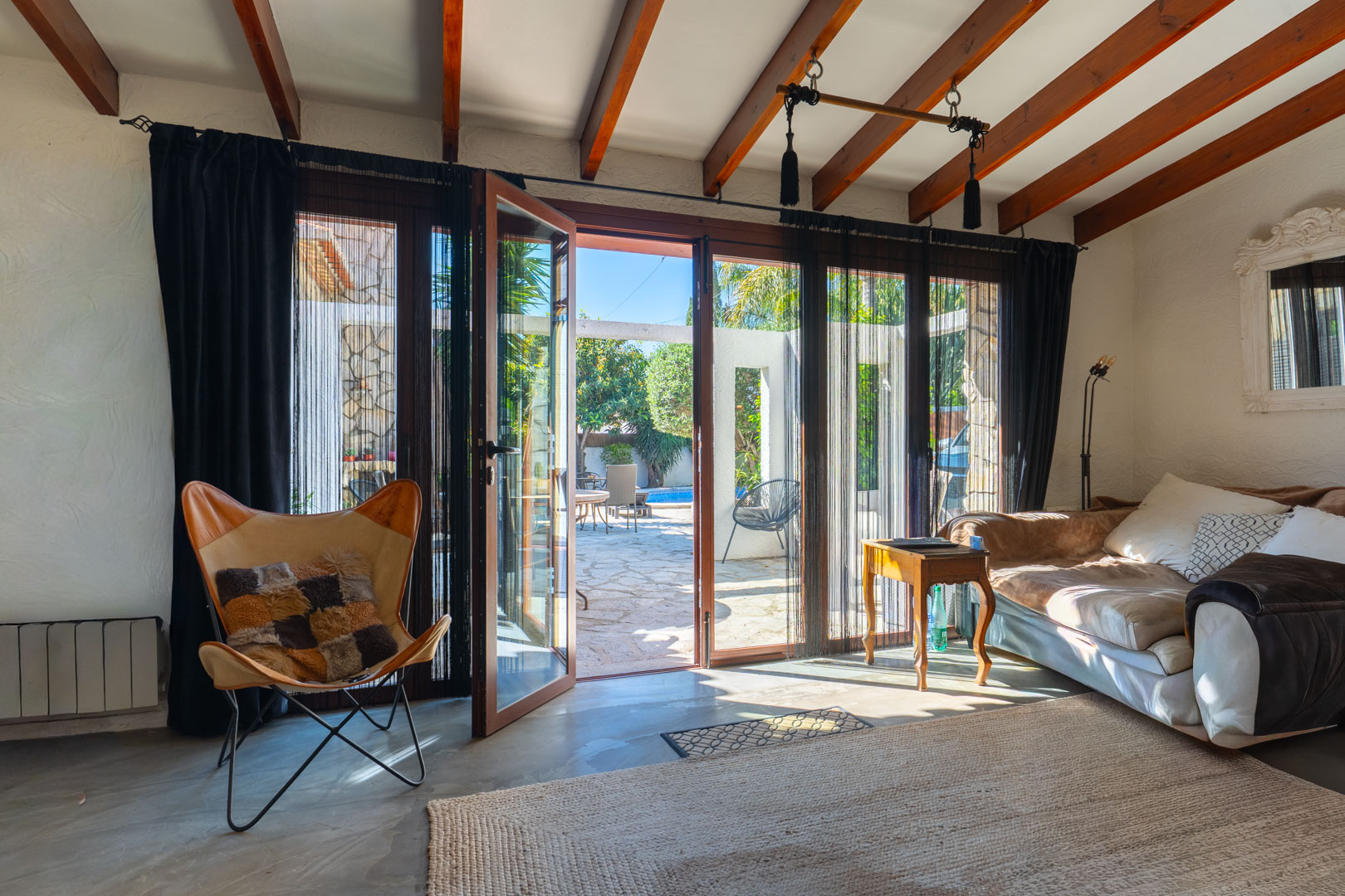 Heller Wohnbereich mit sichtbaren Holzbalkendecken und großen Glastüren, die direkt auf die steingepflasterte Terrasse und den Poolbereich führen.