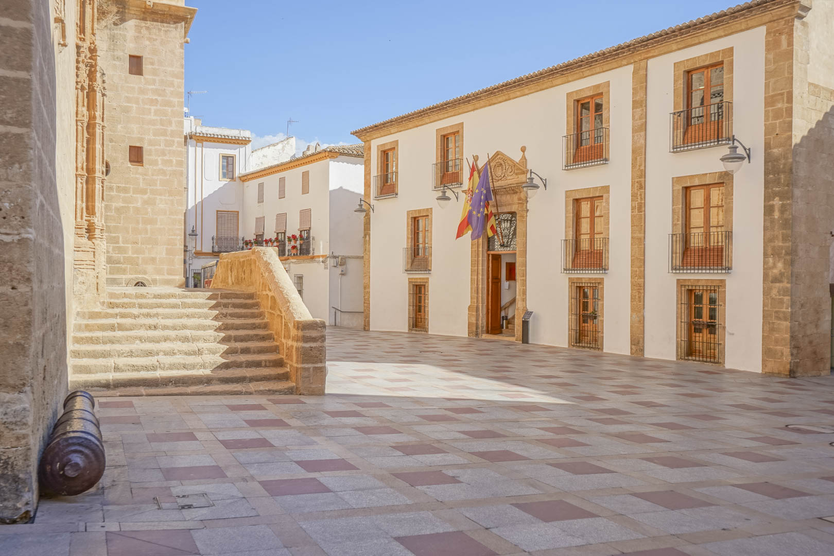 Historischer Stadtplatz in Jávea mit traditioneller Steinarchitektur, dem Rathaus mit Flaggen und der befestigten Kirche San Bartolomé.