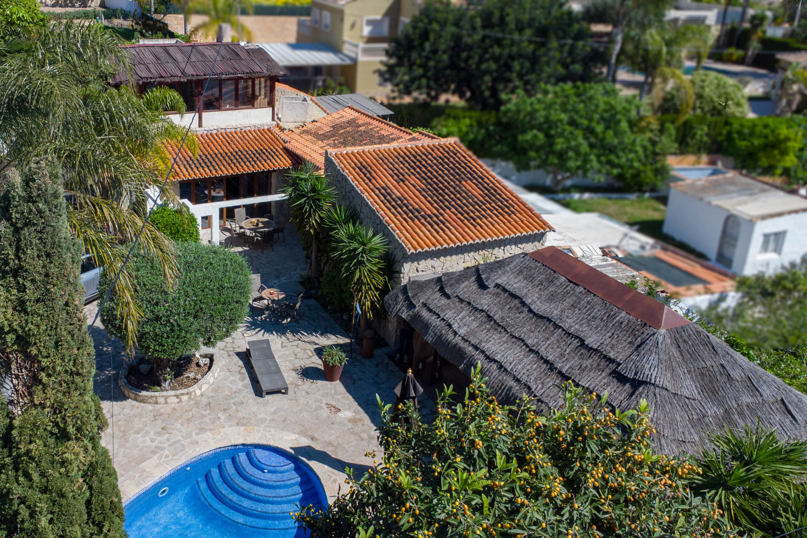 Luftaufnahme der rustikalen Villa mit Terrakottadächern, kreisförmigem blauem Pool, weitläufigen Stein-Terrassen und einer großen balinesischen Pergola mit Reetdach.