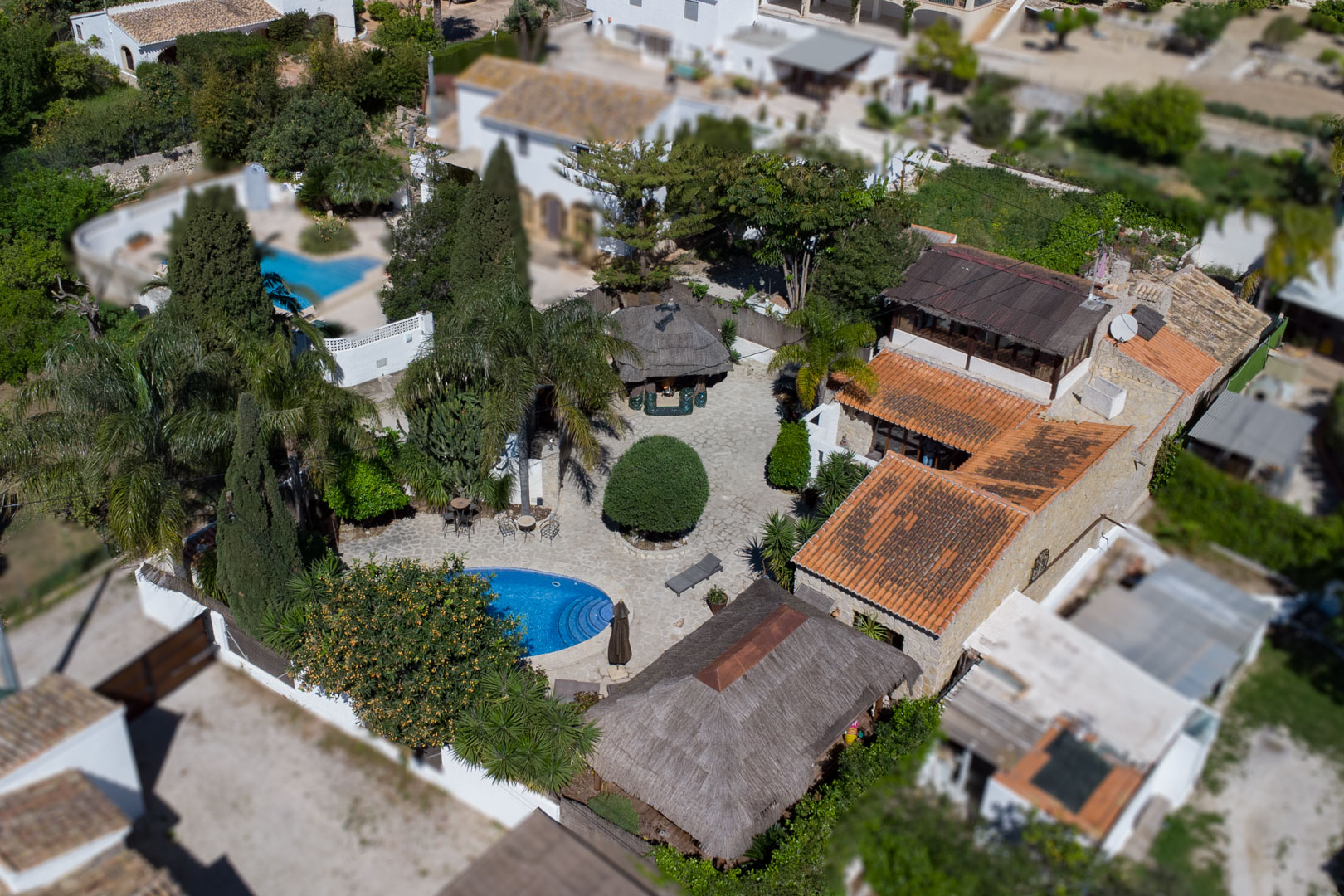 Luftbild der Anlage mit ovalem Pool, Naturstein-Terrasse und balinesischen Pergolen, eingebettet in einen Garten mit mediterranem Baumbestand.