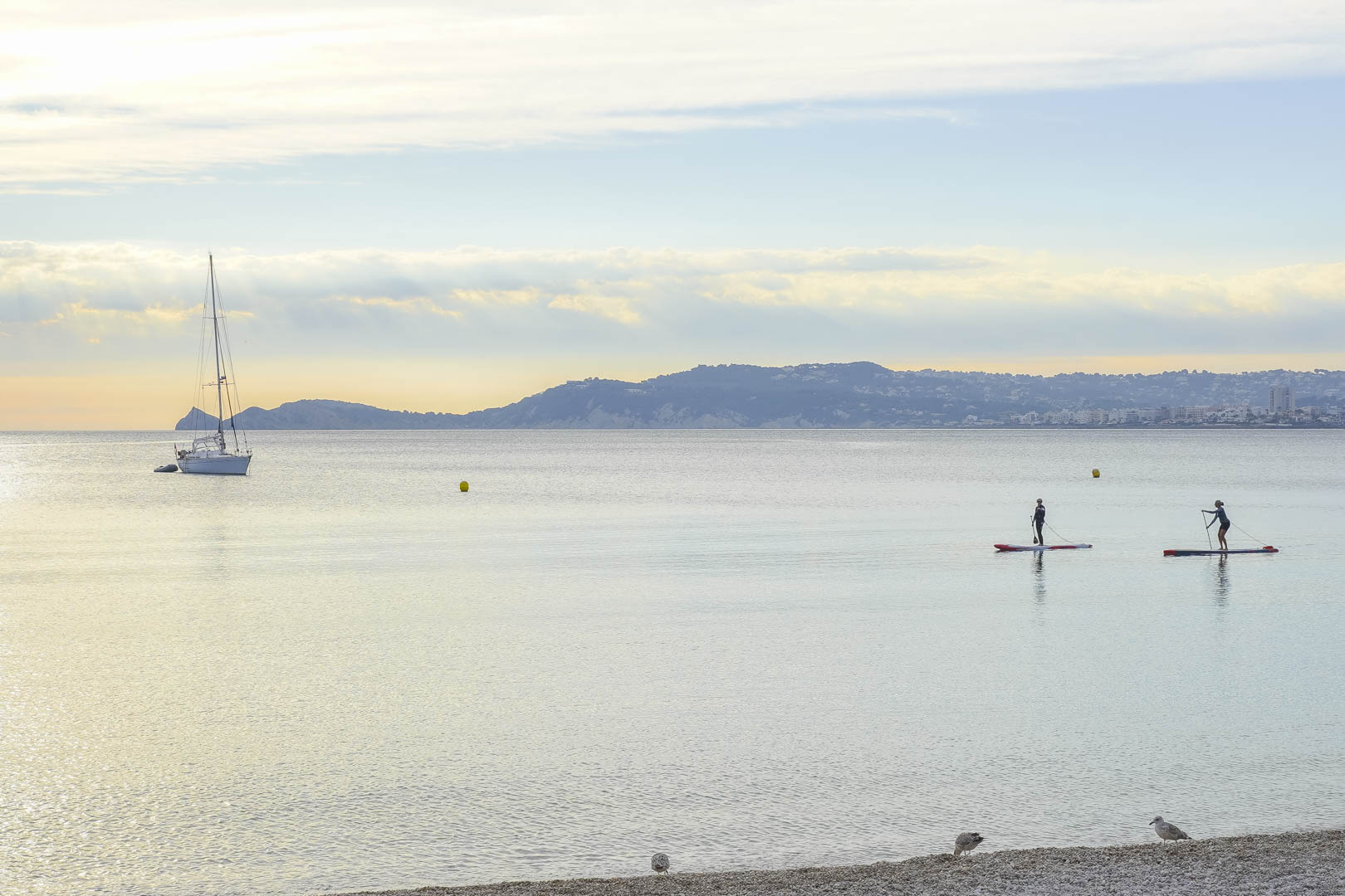 Panoramablick auf die ruhige Bucht mit einem ankernden Segelboot, Stand-Up-Paddlern und der malerischen Küstenlinie von Jávea.
