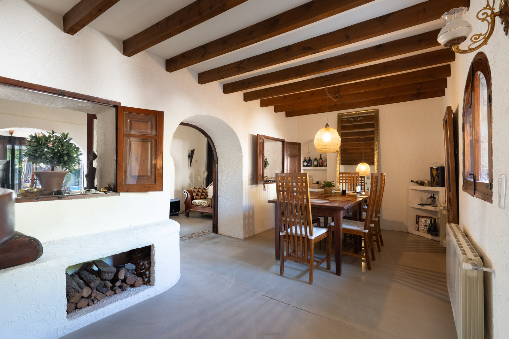 Rustikaler Essbereich mit sichtbaren Deckenbalken, Massivholztisch und Rundbogen. Ein integriertes Holzfach und Innenfenster mit Holzläden unterstreichen den mediterranen Stil.