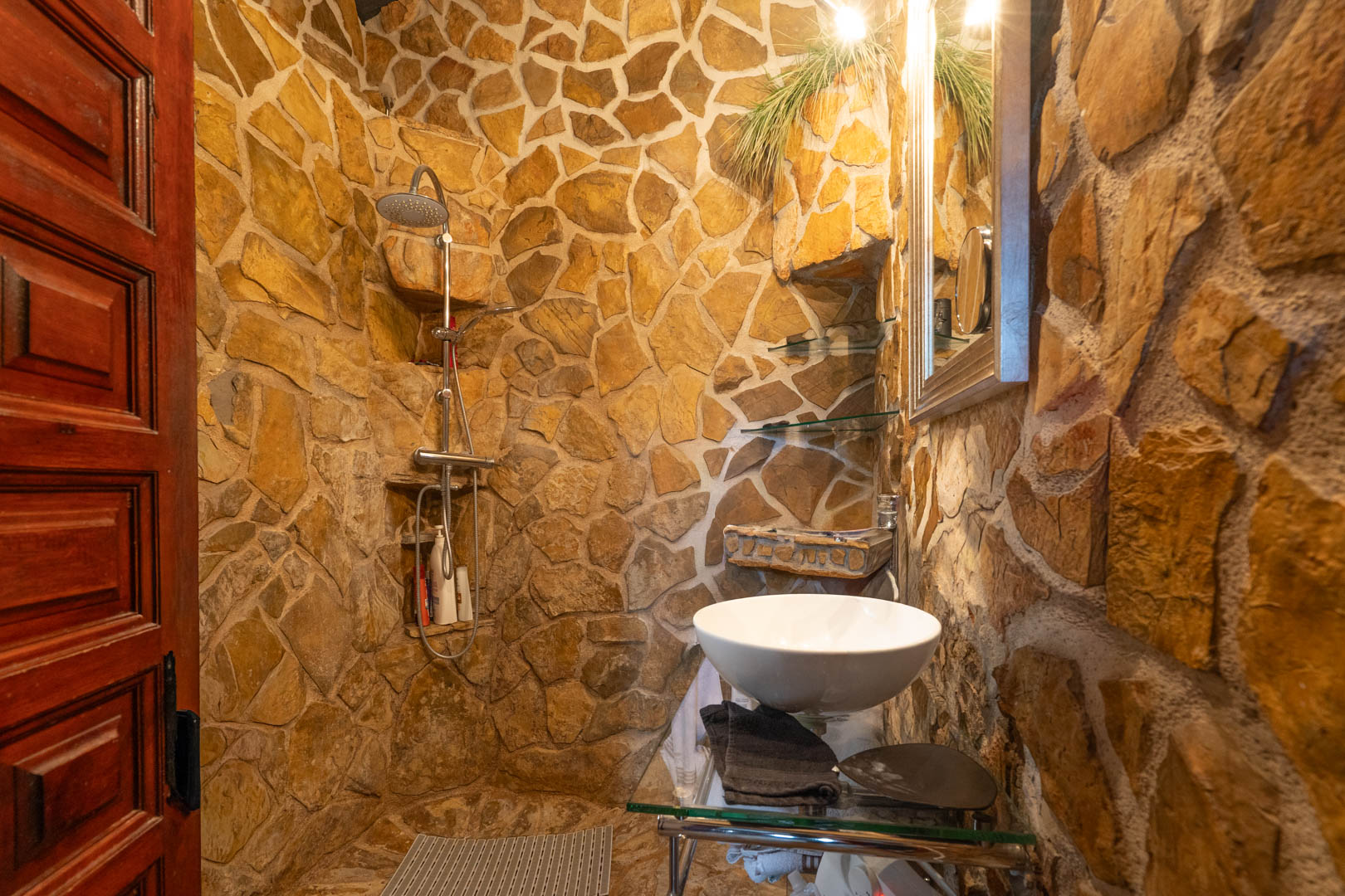 Rustikales Badezimmer mit Natursteinwänden, begehbarer Dusche mit integrierten Ablagen und einem Aufsatzwaschbecken auf einem Glaswaschtisch.